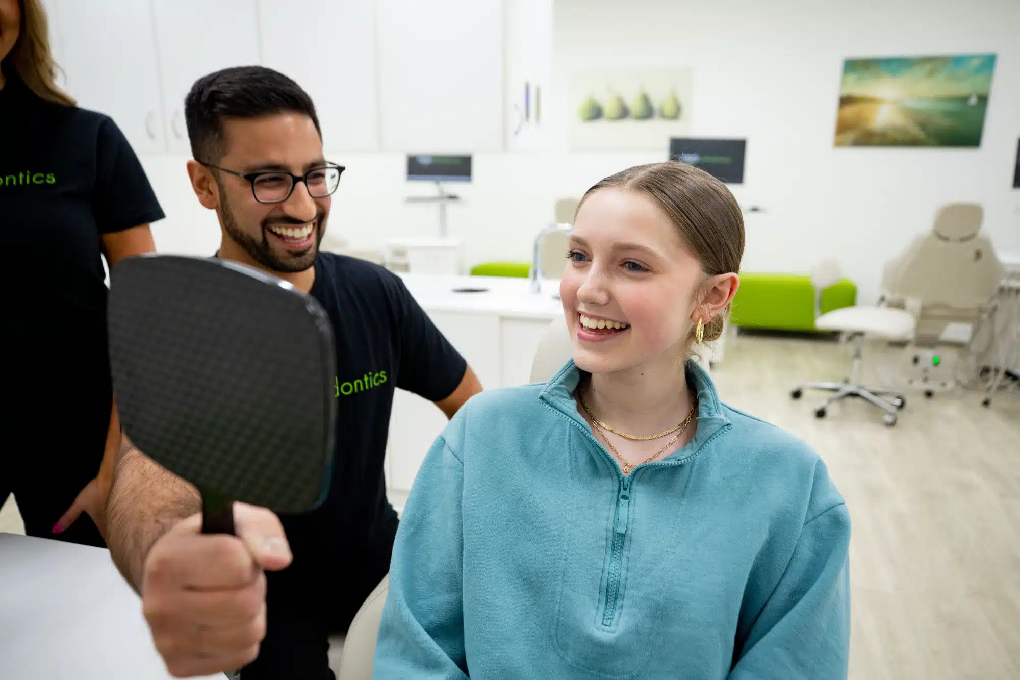Smiling girl checking her Invisalign smile in a mirror held by orthodontist